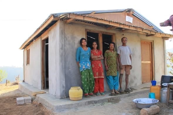 Four adults stand on the concrete apron of a newly plastered house, a yellow water drum beside them.