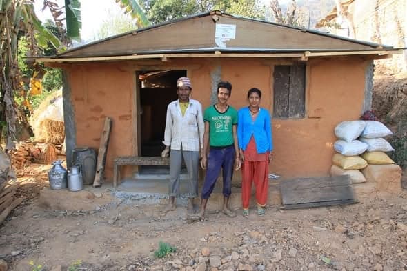 Three young adults stand in the doorway of a mud house with sacks of grain stacked against the wall.