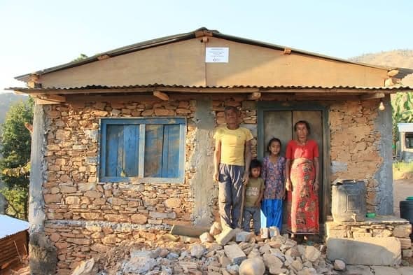 A man, a woman and two children stand in front of a house with dry-stone walls and a blue-framed window.