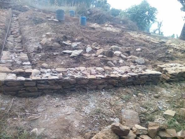 A stone retaining wall and levelled platform cut into a slope, with blue water drums standing above it.