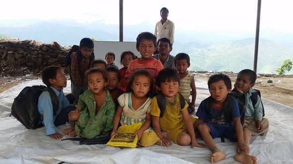 About fifteen small children sit on a tarpaulin under an open shelter, a man standing behind them and mountains beyond.