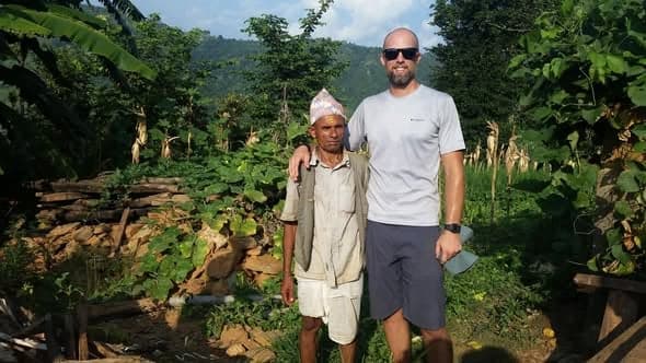 A volunteer in sunglasses stands with his arm around an older man in a topi, among maize plants and stacked firewood.