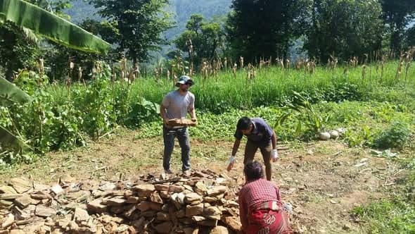 Three people sort and stack flat building stone in a clearing beside banana plants.
