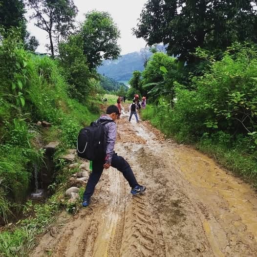 A student with a rucksack steps across a stream running over a deeply rutted, muddy hill road, others walking ahead.