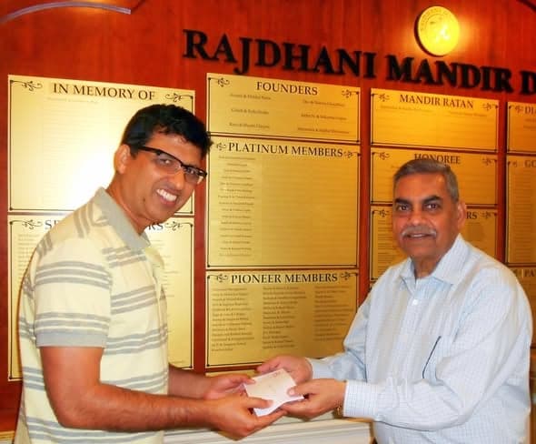 Two men shaking hands over a cheque in front of a wall of engraved donor plaques headed Rajdhani Mandir
