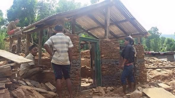 Men rebuild the stone walls of a house around a green-painted door frame, timber and rubble around them.