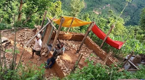 Two men dig foundations on a steep terrace, shaded by yellow and red tarpaulins strung on poles.