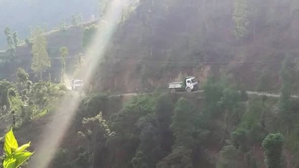 Two trucks make their way along a narrow road cut into a forested hillside in hazy light.