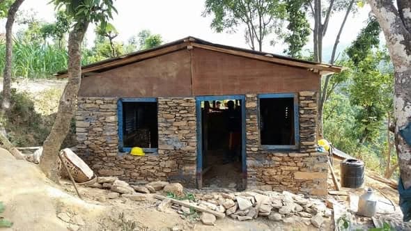 A completed single-room transition home with dry stone walls, blue-painted window frames and a corrugated roof on a hillside terrace