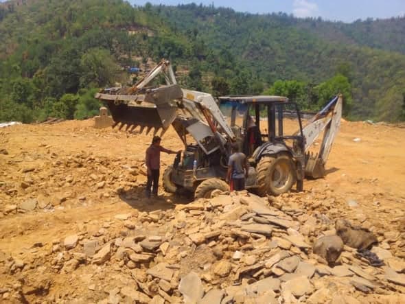 A backhoe loader stands on a cleared, rubble-strewn site with two men beside its engine.