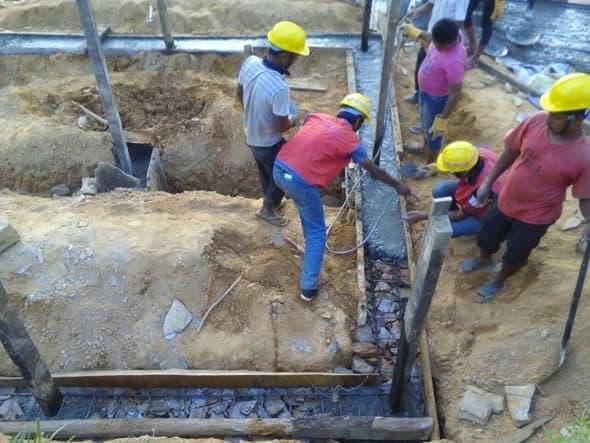 Workers in yellow helmets lay reinforcement and stone into trench foundations.
