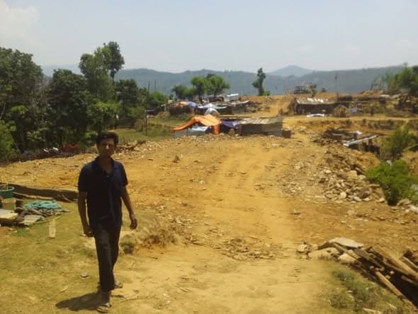 A young man walks along a wide dirt track past tarpaulin shelters and salvaged timber on a cleared hilltop.