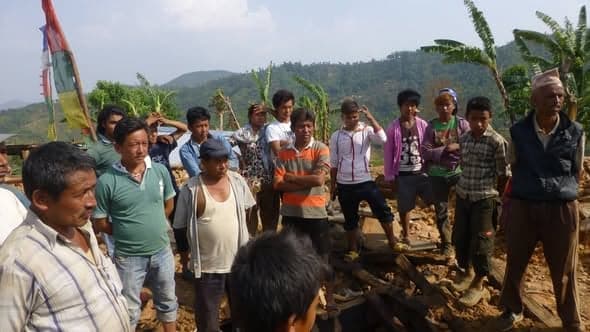 A crowd of village men and boys stand among the rubble of collapsed stone houses, prayer flags behind them.