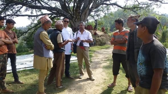 A group of men talk in a circle under a large tree beside a village path, one of them holding a notebook.