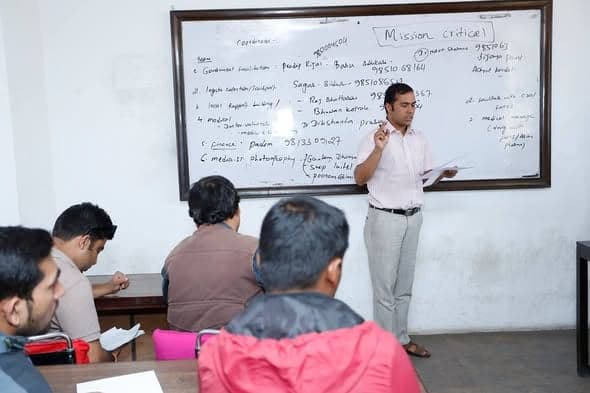 A man writes on a whiteboard headed Mission Critical while a dozen people take notes at desks in a bare room.