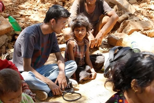 A young volunteer with a blood pressure cuff sits on a mat among rubble, talking with a small girl and an older woman.