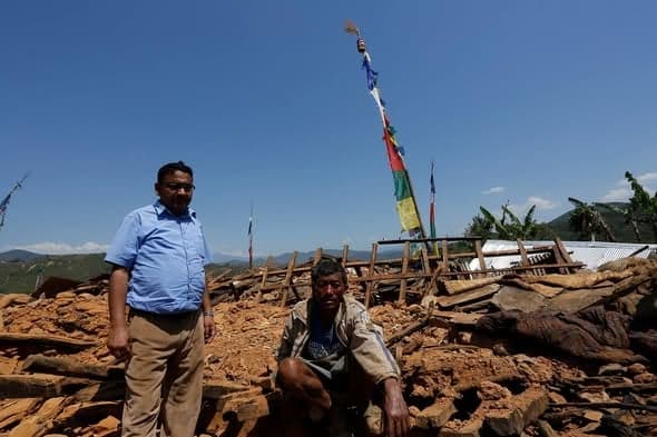 A man crouches on a mound of stone rubble beside a standing visitor, prayer flags on a pole behind them and mountains in the distance.