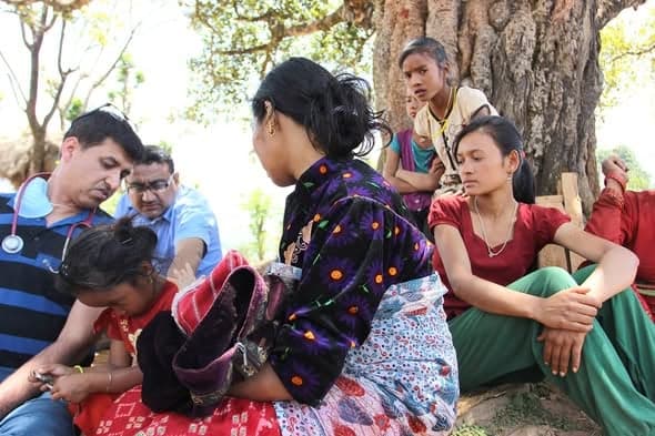 A doctor with a stethoscope examines a child lying across a woman's lap, on a mat under a tree, with other villagers waiting beside them.