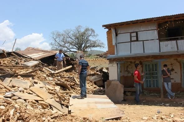 Men standing among the collapsed brick and timber remains of houses, beside a two-storey building with its ground floor wall torn away.