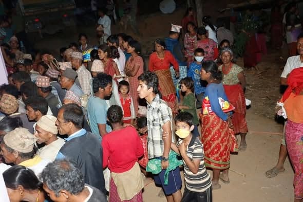 A large crowd of villagers, many carrying bundles, waiting in the dark at a relief distribution point.