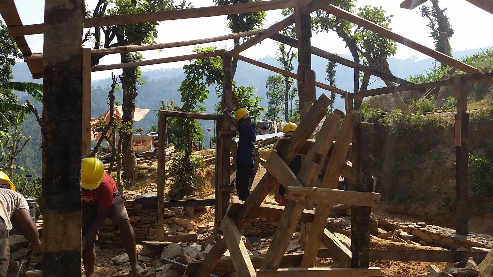 Workers in hard hats raise a timber frame on a hillside beside stacks of salvaged timber and stone.