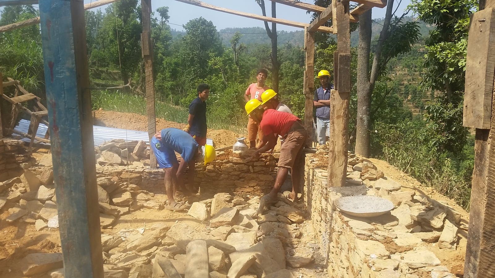 Workers in hard hats laying flat stones into a low wall between timber posts, with cut stone stacked around them.