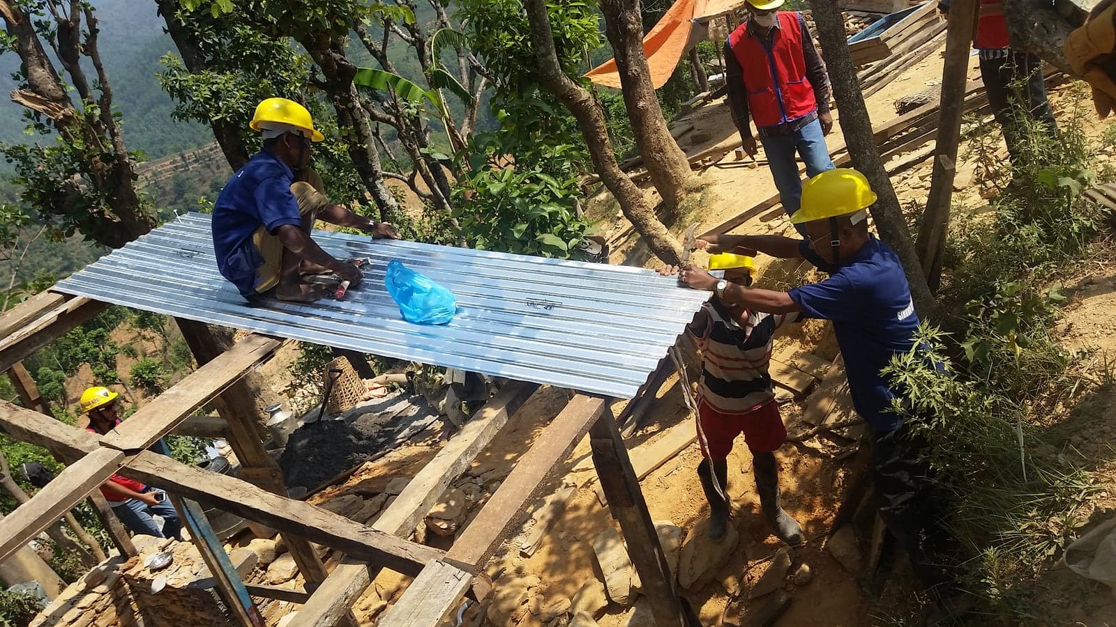 Three men in hard hats nailing down a sheet of corrugated zinc across roof timbers on a steep hillside, with a valley falling away behind them.