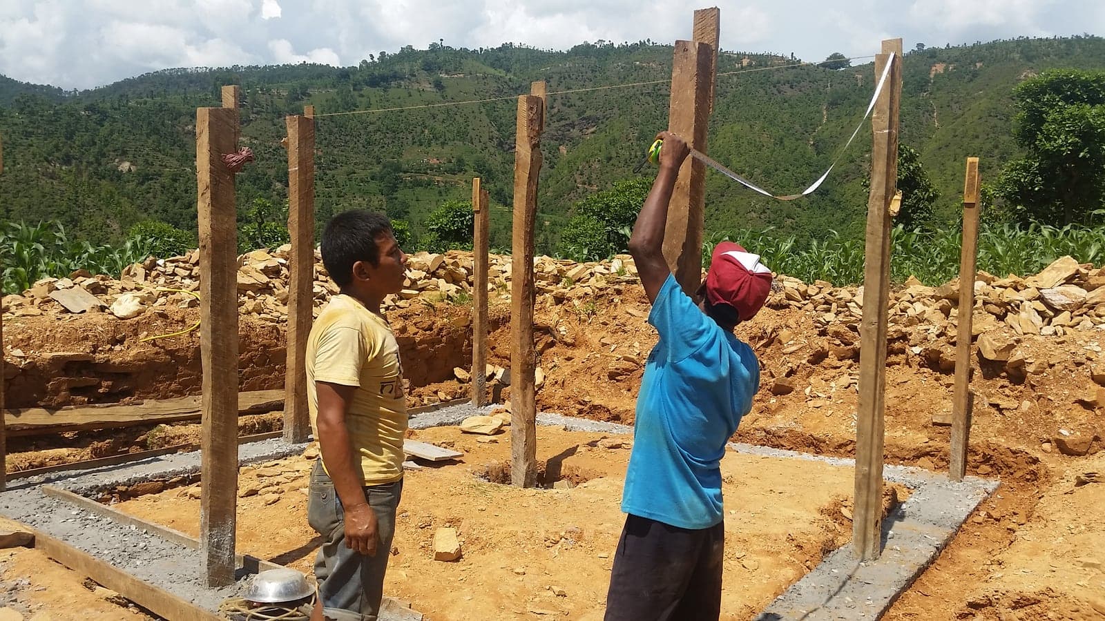 Two men setting out timber posts on freshly poured concrete footings, one measuring a post with a tape while a string line runs across the plot.