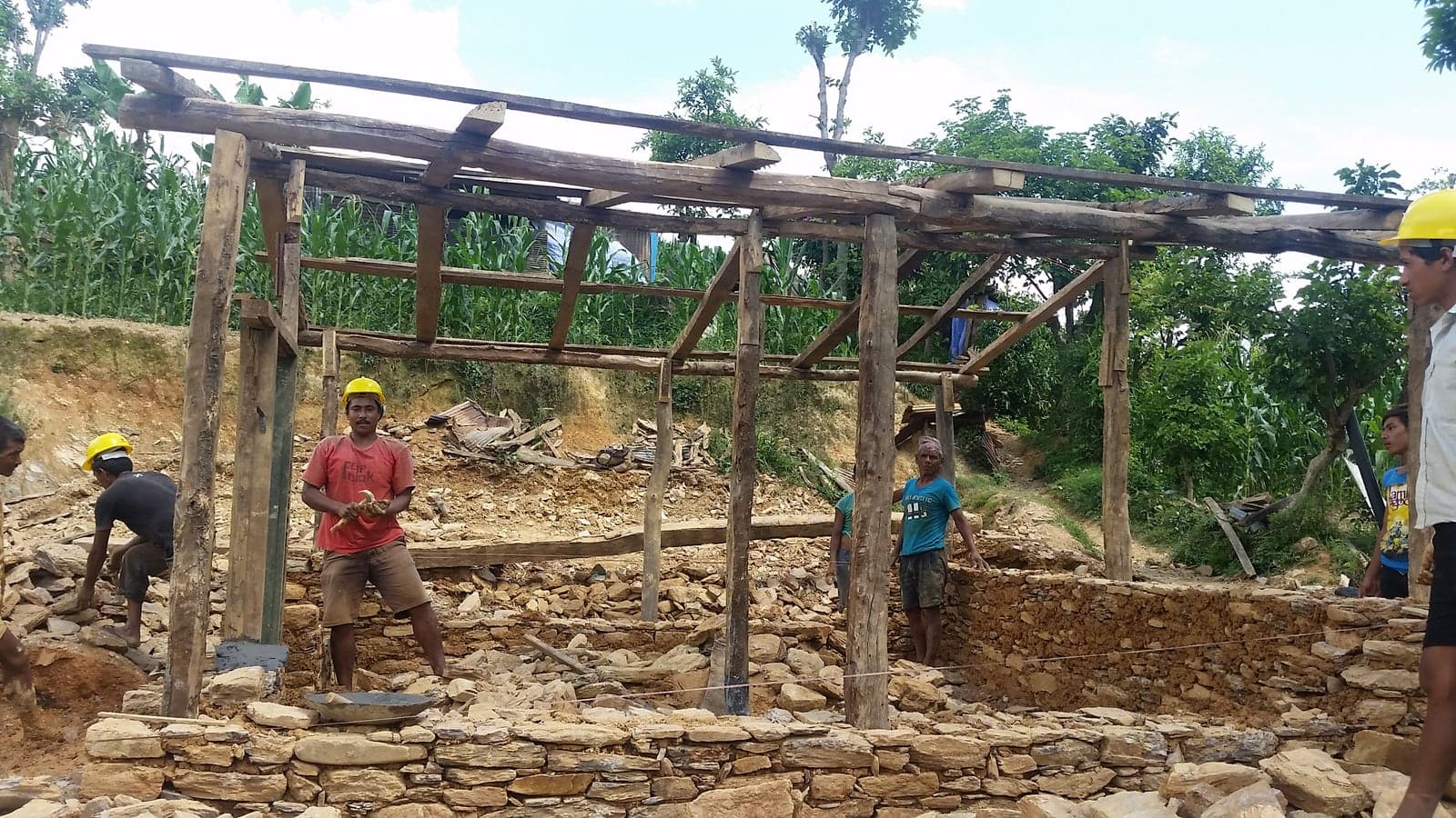 Workers in yellow hard hats raising a timber frame over a dry-stone plinth on a cleared hillside plot, with rubble from the previous house heaped behind.