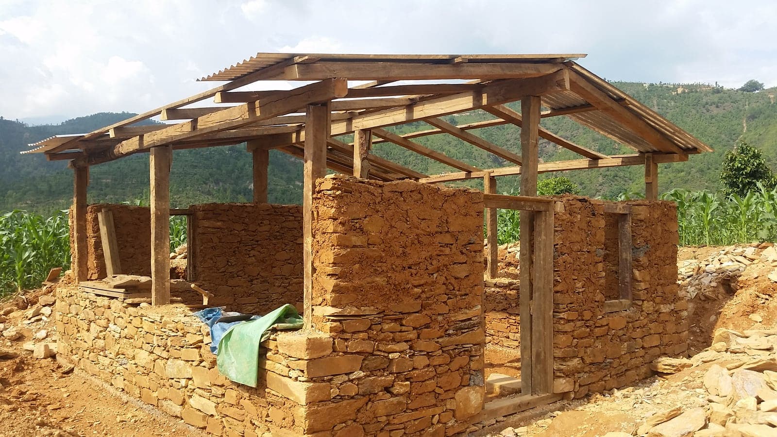 A transition home under construction on a terraced hillside: a heavy timber frame carrying a corrugated zinc roof over dry-stone and mud walls, with maize growing behind it.