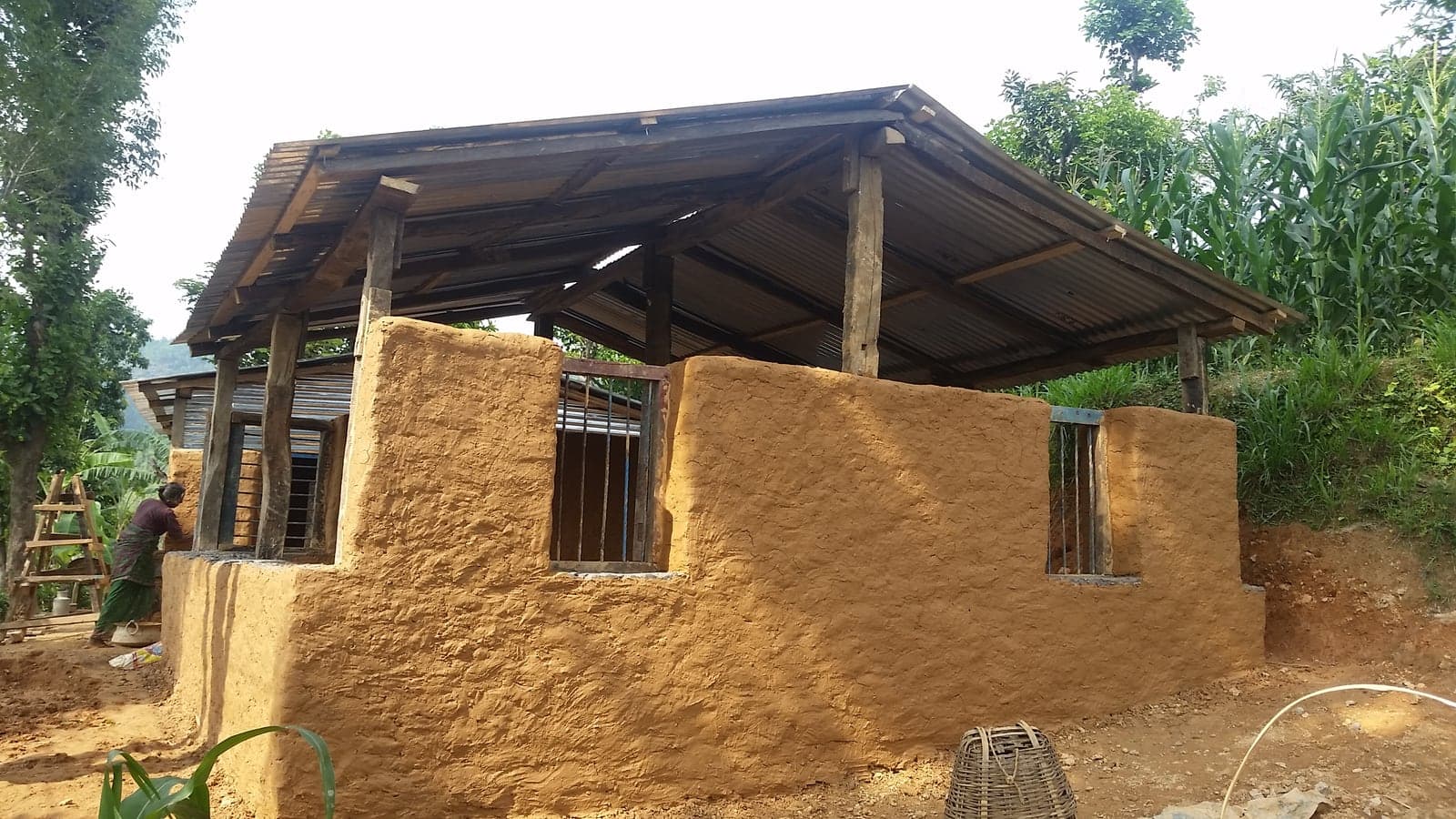 A finished transition home with mud-plastered walls, barred window openings and a corrugated roof, a woman working at the far end of it.