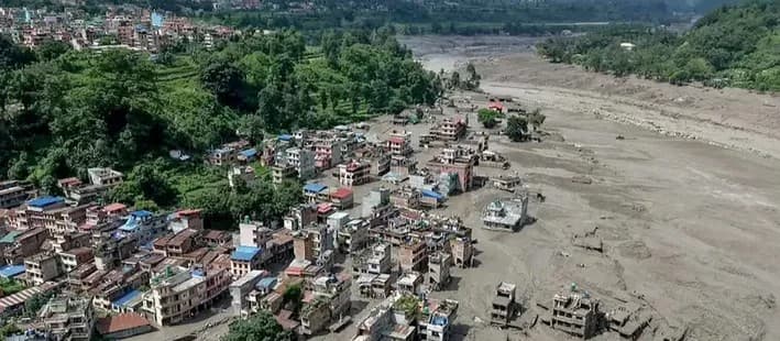 Aerial view of a town in the Trishuli corridor after the flood: streets and buildings on the left, a wide grey field of mud and debris where the river has spread across the valley floor on the right.