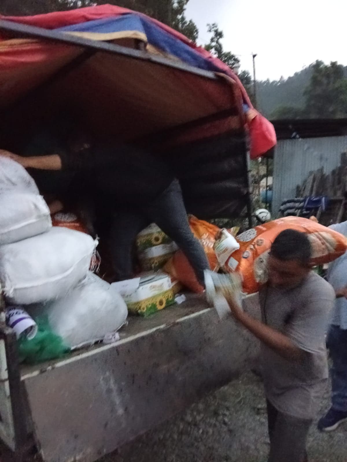 A man lifting a sack of rice down from the tailgate of a loaded truck at dusk, with bales of bedding and cartons stacked behind him.