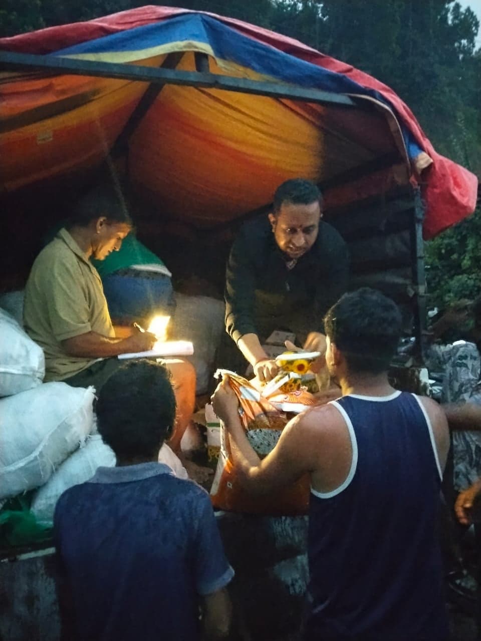 Dusk at the back of a tarpaulin-covered truck: one man writes on a clipboard by torchlight while another passes a bag of supplies down to the people waiting below.