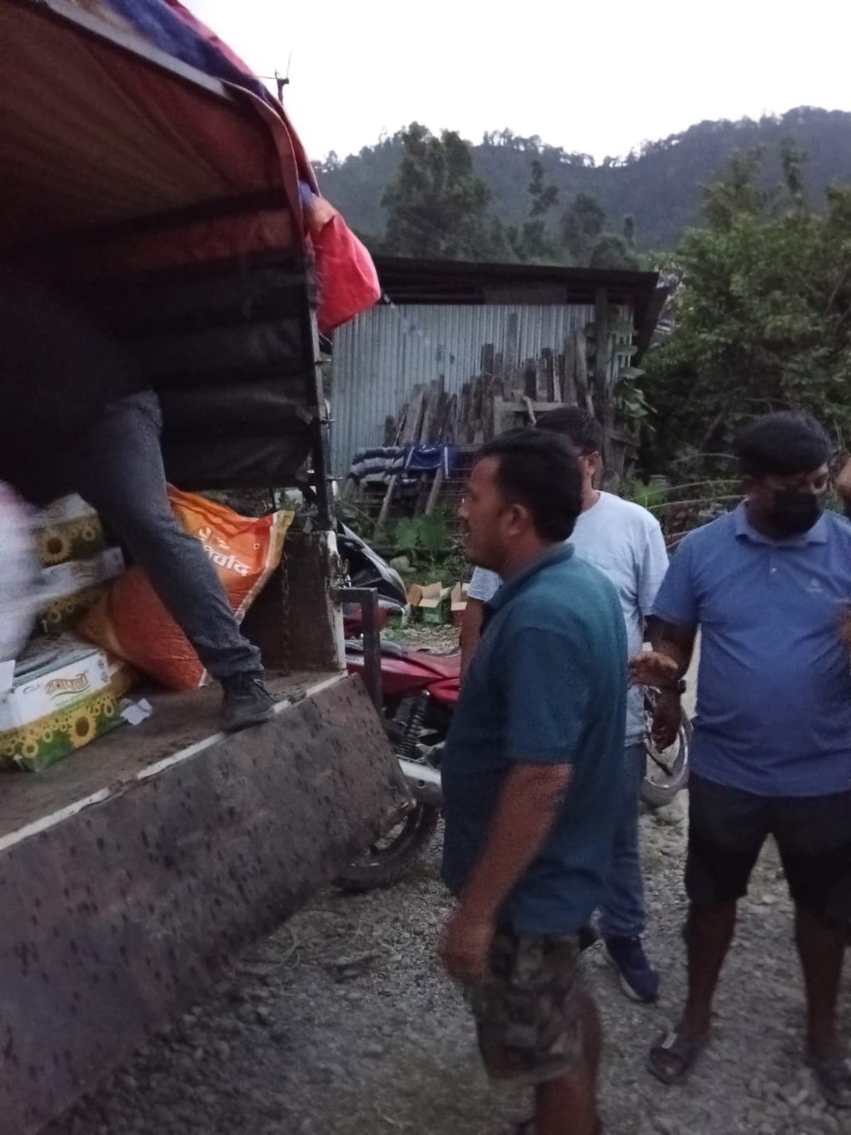The side of a loaded truck beside a corrugated-iron shed, sacks and cartons stacked on the bed and men standing on the track alongside.