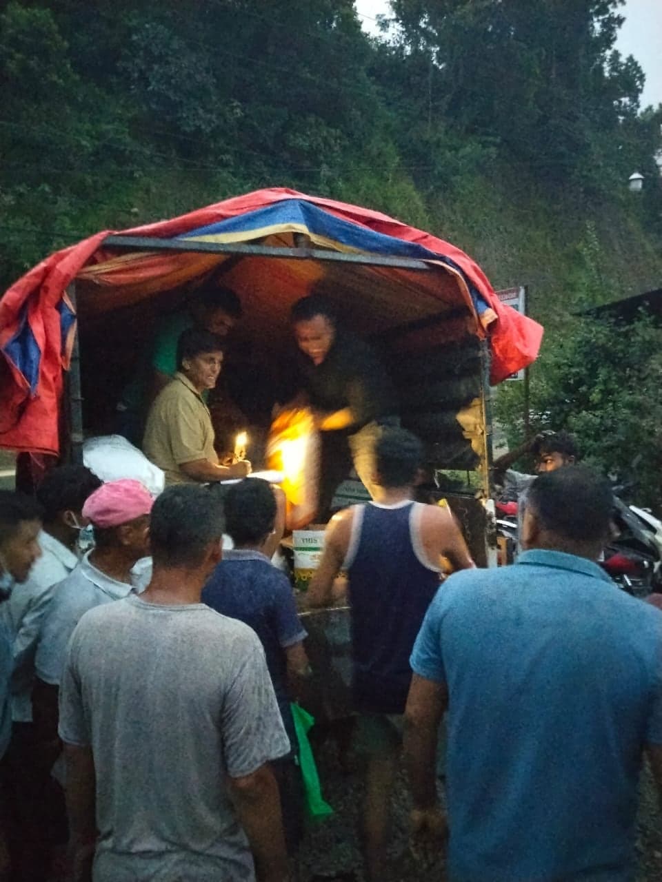 A crowd standing at the back of a truck at dusk. Inside the bed one man checks a list by lamplight while another hands a bag down.