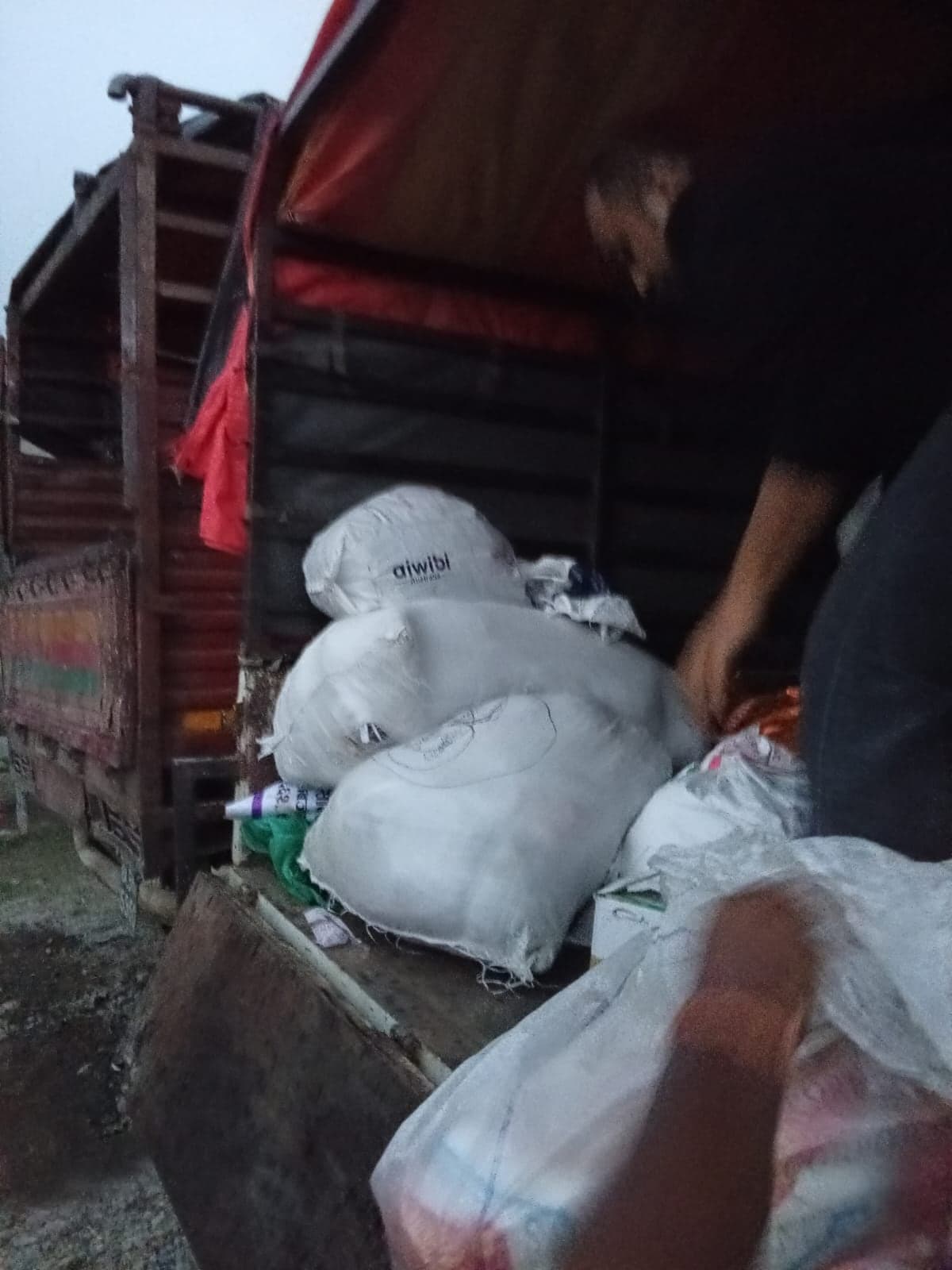Bales of bedding and sacks stacked on the open tailgate of a truck, with hands reaching in from the right to lift one down.