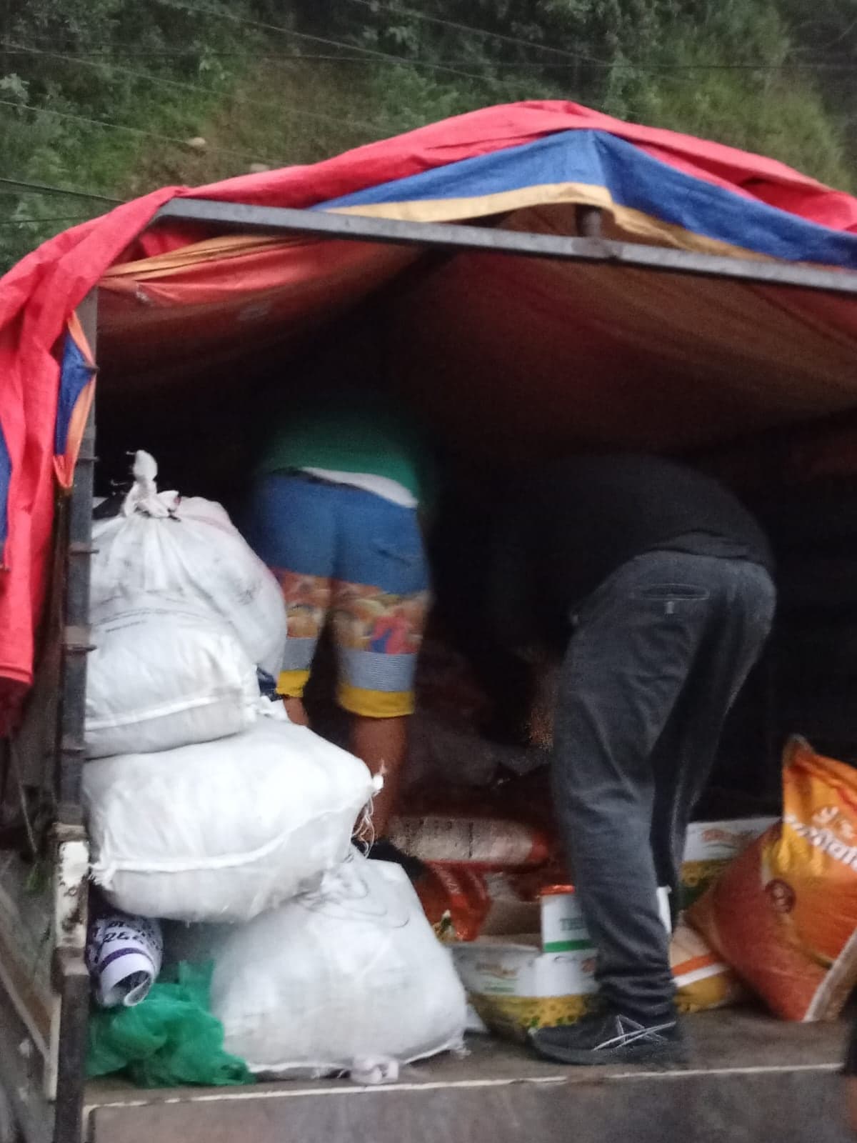 Looking into the bed of a truck under a red and blue tarpaulin: white sacks of bedding, orange sacks of rice, tins of cooking oil, and two people working at the far end.
