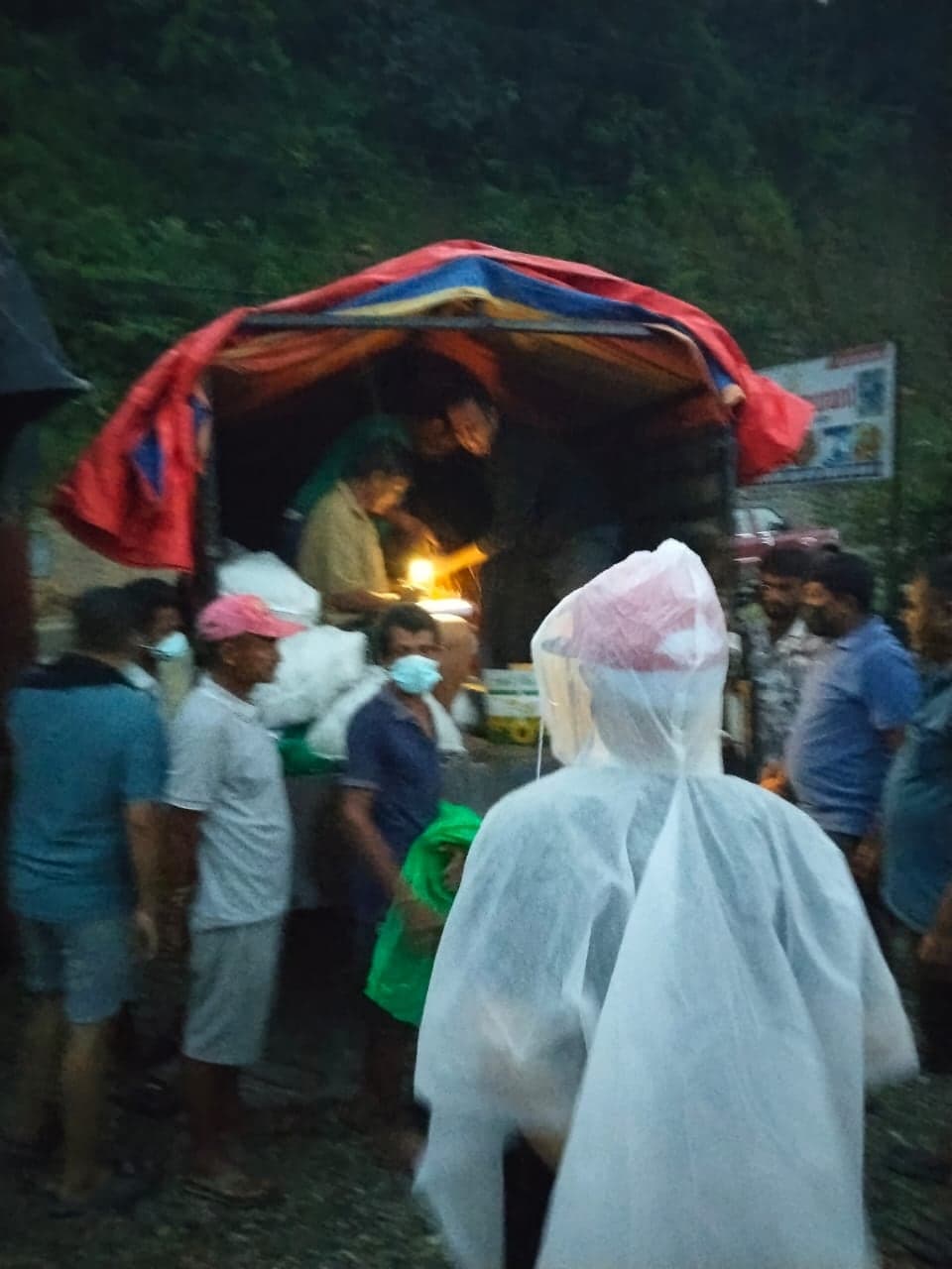 People in plastic rain capes queuing at the tailgate of a truck at dusk while supplies are handed out by lamplight.