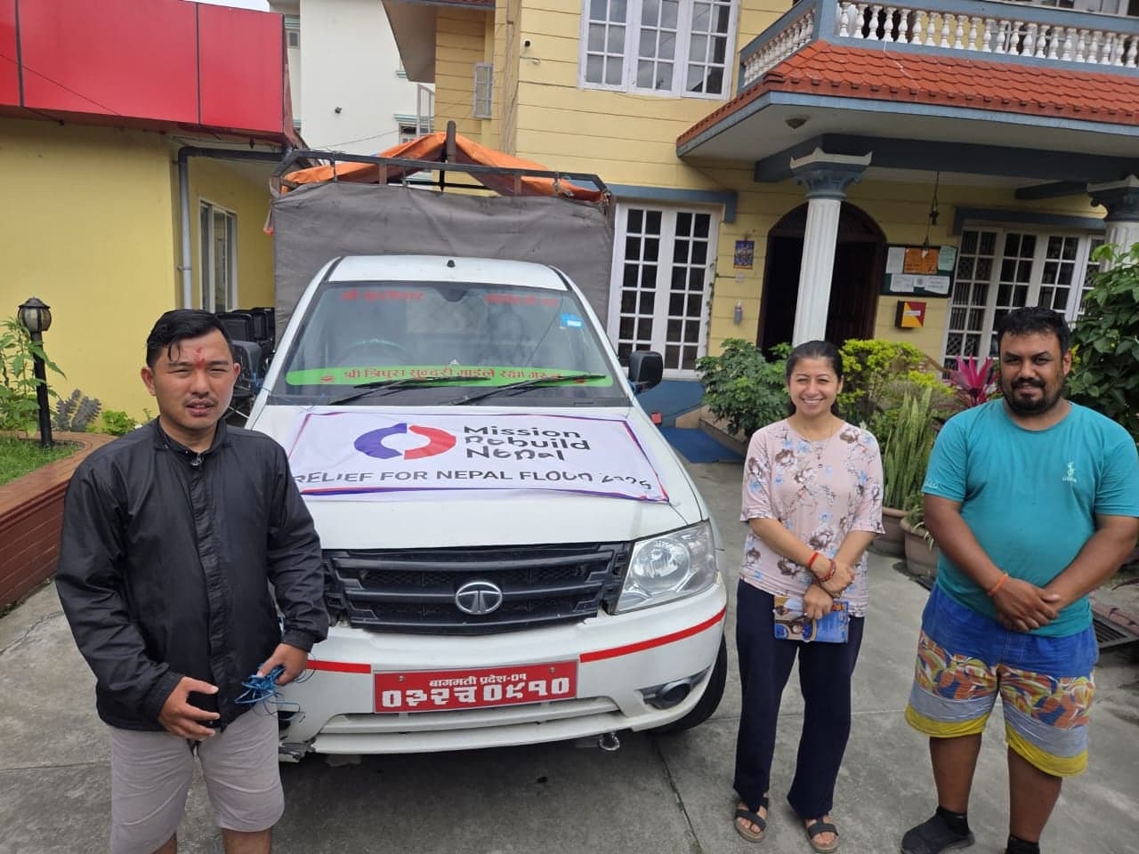 Three people standing beside a white pickup truck with a tarpaulin frame over its bed and a banner taped across the bonnet reading Mission Rebuild Nepal, Relief for Nepal Flood 2026.