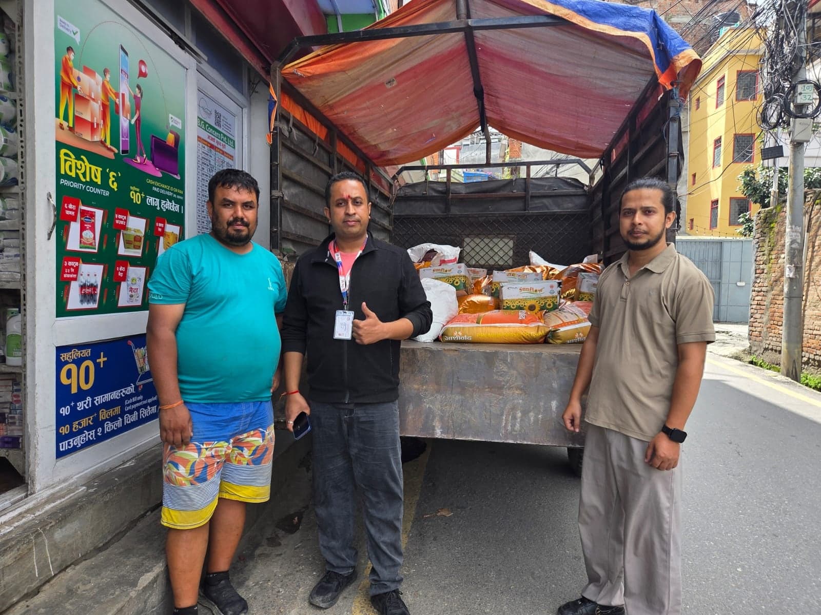 Three men at the open back of a truck loaded with sacks of rice and cartons of cooking oil, in a narrow street outside a wholesale shop.