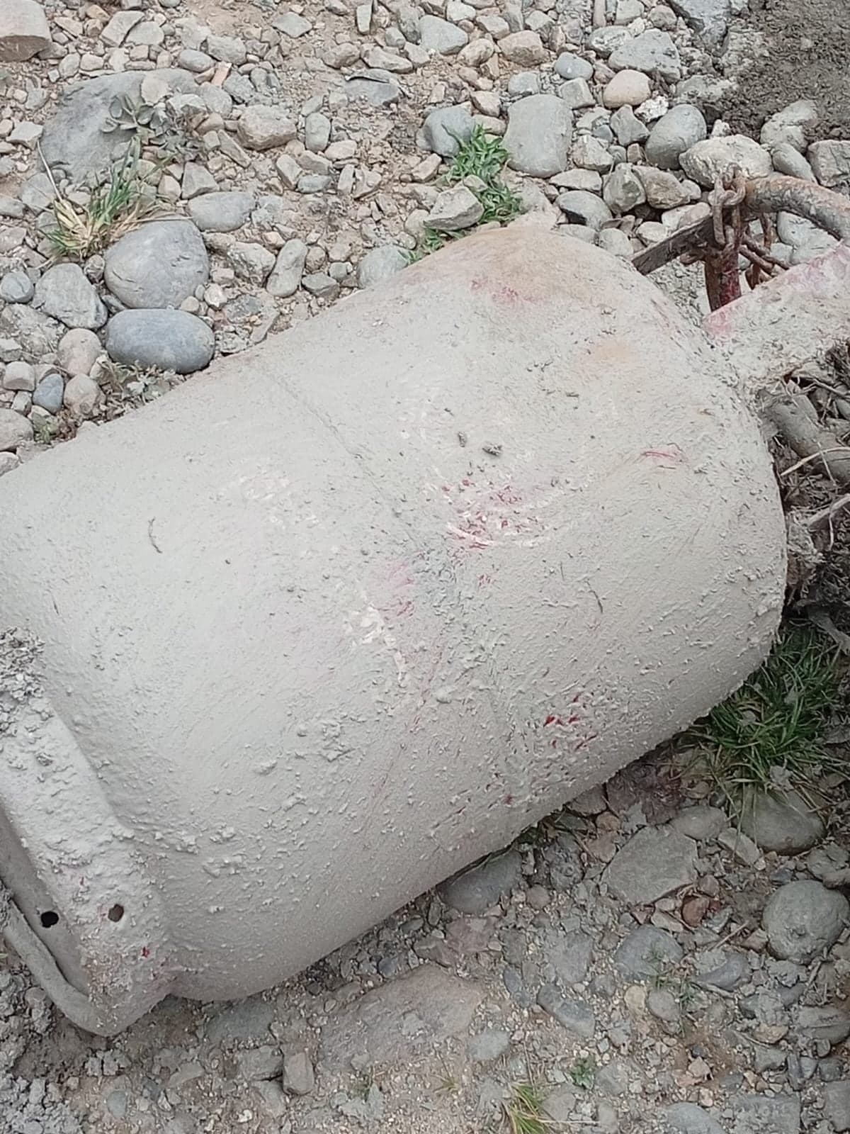 A gas cylinder lying among river cobbles, coated to the valve in dried grey silt.