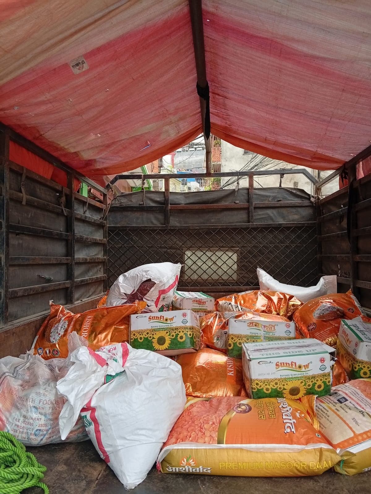 The bed of a truck loaded with sacks of lentils and rice and cartons of sunflower cooking oil under a red tarpaulin.