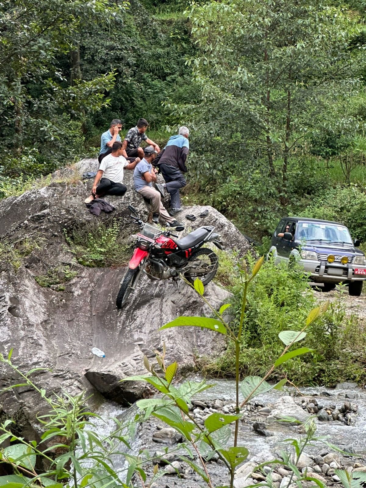 Men sitting on a rock above a stream where the road crosses it, a motorcycle parked on the rock beside them and a jeep waiting on the far side.
