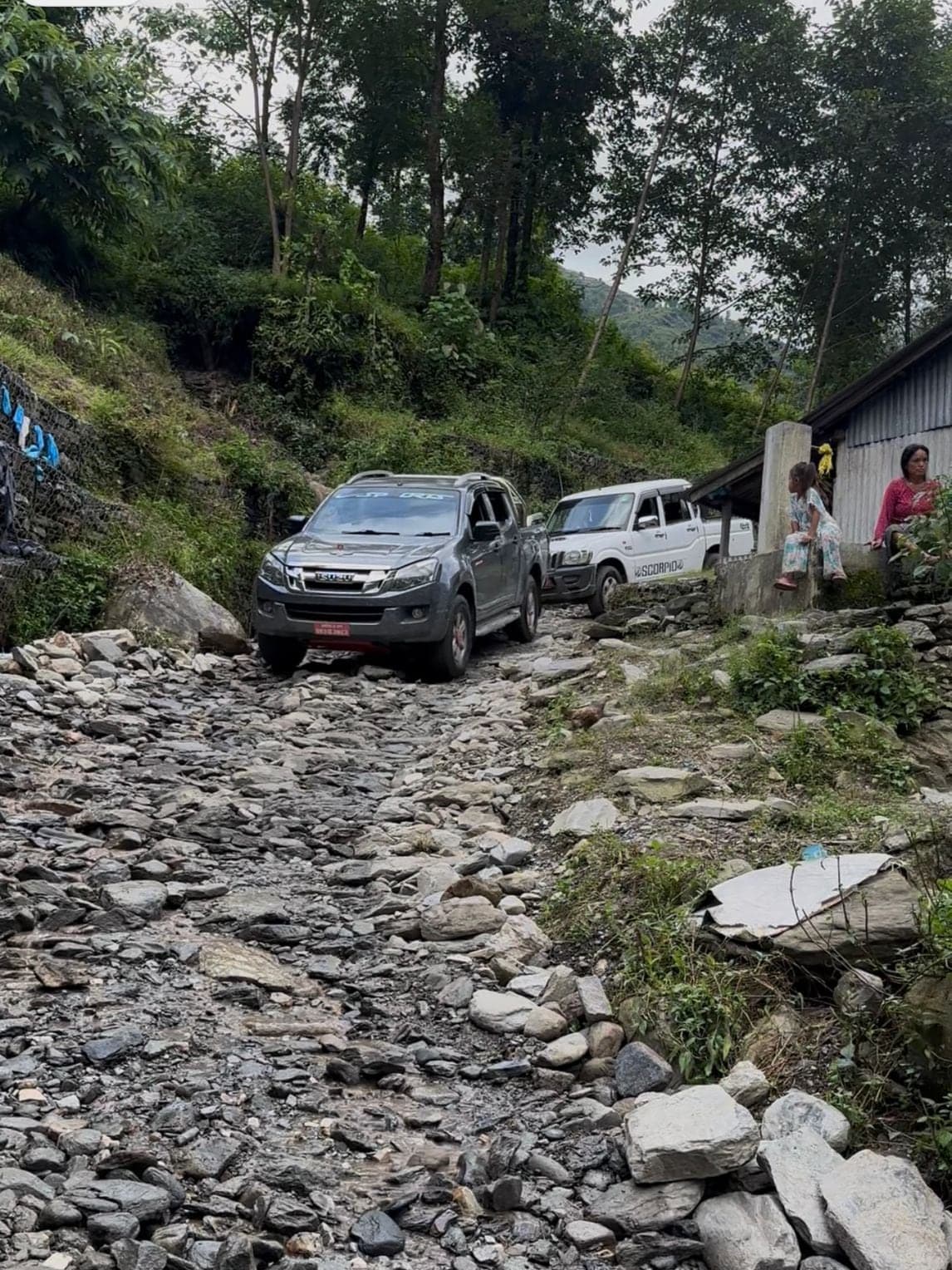 Two pickup trucks picking their way along a hill road buried under boulders and river stone, with a woman and a small child sitting on a wall beside a tin-roofed house.