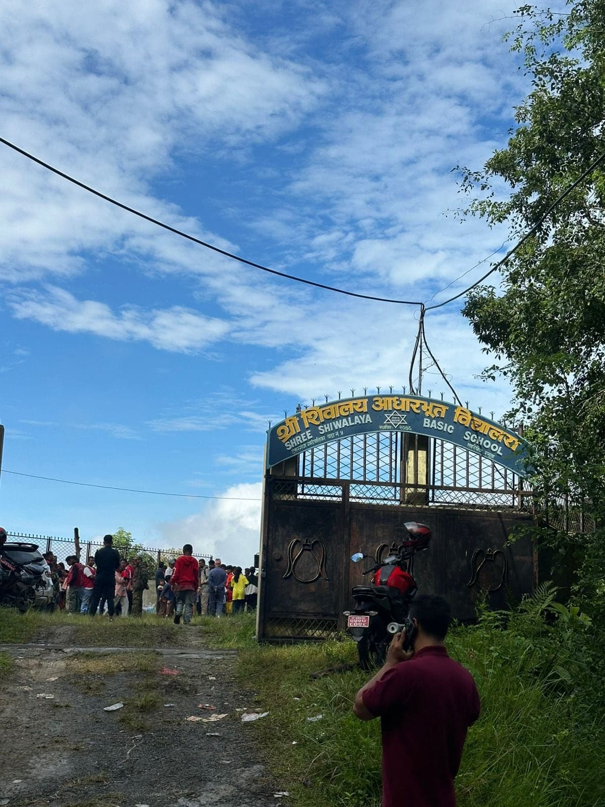 The gate of Shree Shiwalaya Basic School under a bright sky, with a crowd of people gathered on the ground inside.