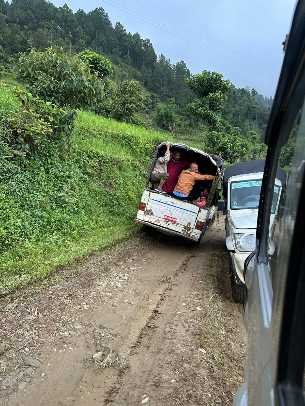 A pickup carrying passengers in its open back, edging along a rutted mud track cut into a green hillside, with another vehicle waiting to pass.