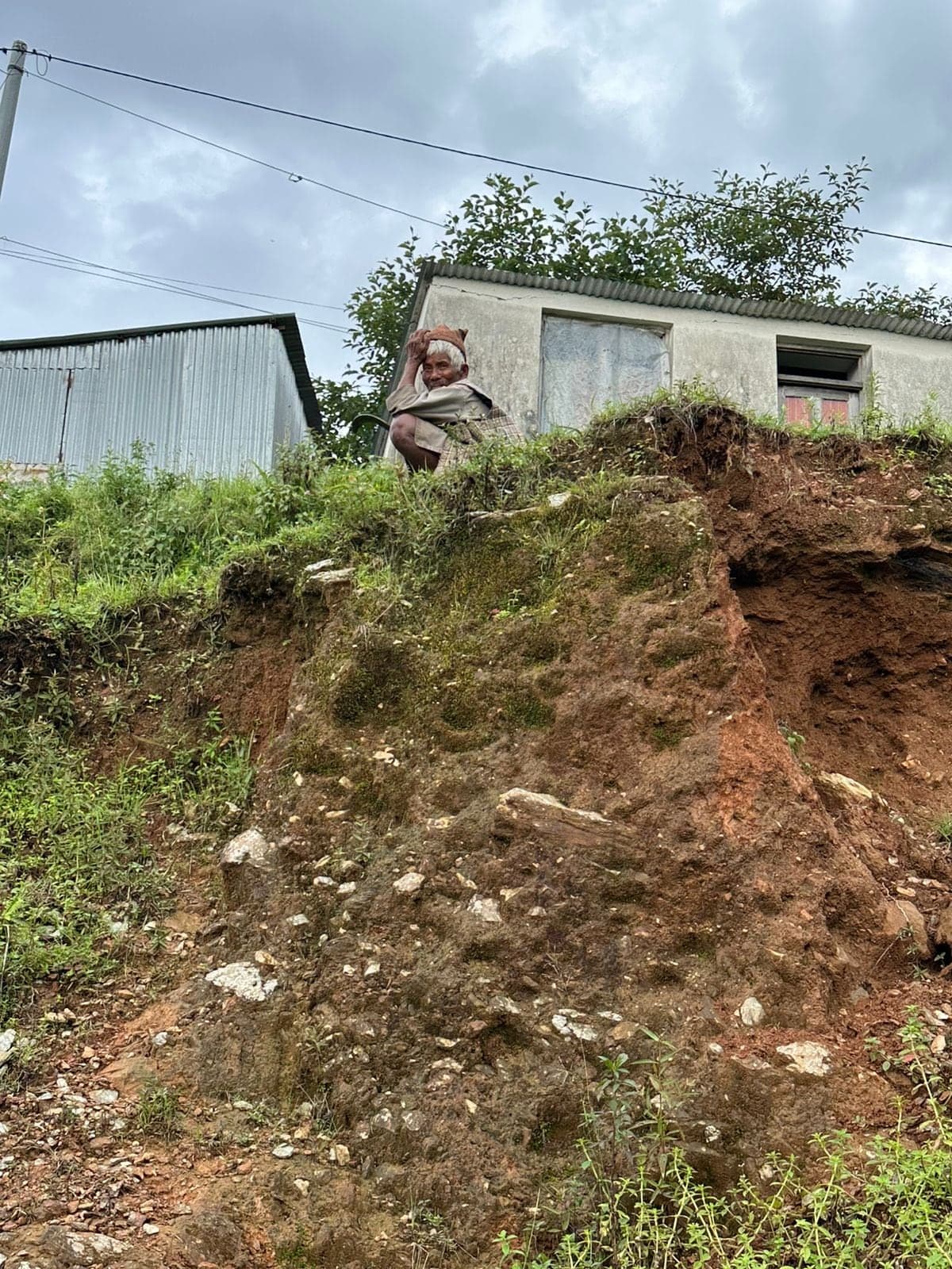 A house standing at the edge of a slope that has been cut away beneath it, leaving a raw earth face several metres high. An elderly man stands at the top beside the wall.