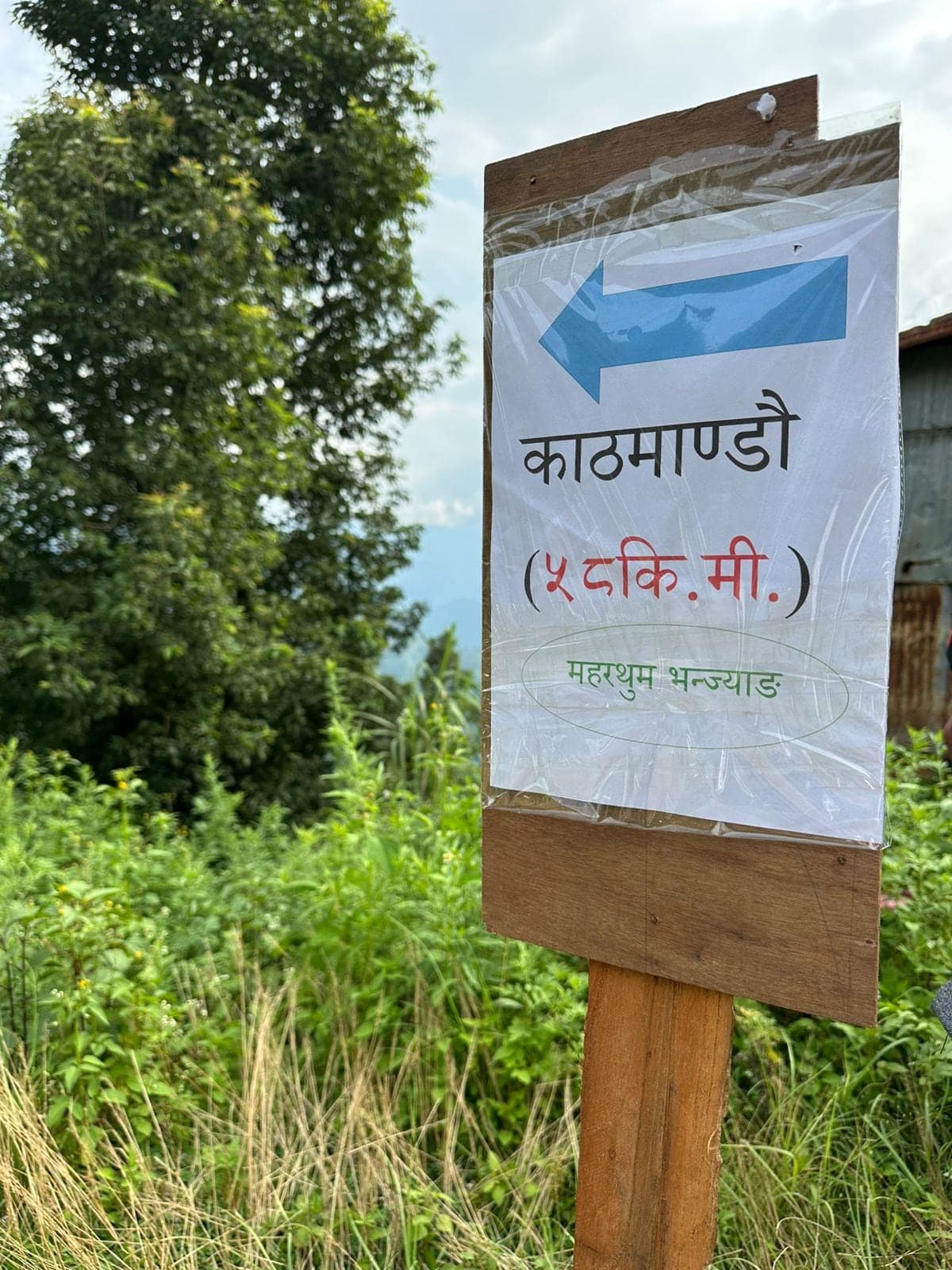A hand-lettered sign nailed to a post beside a hill track: an arrow, Kathmandu, and the distance in kilometres by way of Maharthum Bhanjyang.