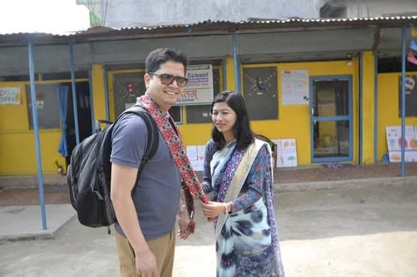 Two people standing in the yard outside a single-storey school block with yellow and blue classrooms and children's drawings on the walls